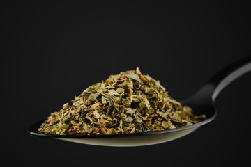 Wall Mural - black spoon with Italian seasoning - dried oregano with thyme, basil and vegetables. black background. closeup