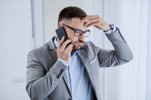 Upset Handsome Classy Unshaven Businessman In Suit And With Eyeglasses Holding His Head And Talking On The Smart Phone While Standing Next To Window In His Office.