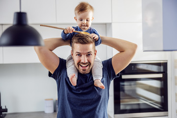 Wall Mural - Young proud smiling positive single dad playing holding his beloved son on shoulders and having fun and kitchen. Toddler holding mixing spoon and playing with it.