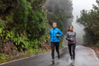 © Maridav - Rain run fit people training jogging in rain weather wearing cold clothing running outdoors in nature autumn season. Active couple on wet park trail jogging. Asian woman, Caucasian man athletes.