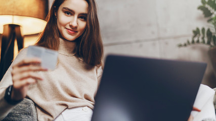 Young female with long hair sitting at home in front of laptop and shopping online. Person making money transfer via Internet to pay for purchases at online retail store.