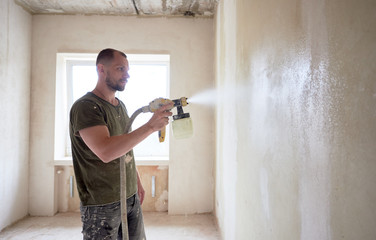 Wall Mural - Concentrated worker painting with airbrush, apartment repairment against small window in the daytime. Man with a beard is dressed in paint-smeared T-shirt. Repair concept