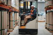 © StratfordProductions - Smiling forklift driver wearing white helmet and vest sitting in machine transporting goods