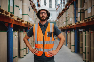 Wall Mural - Confident warehouse manager standing in between shelf with hands on waist wearing uniform and hardhat inspecting goods delivery process