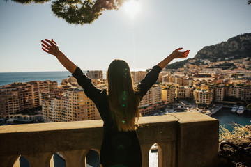 Wall Mural - Back view of cheerful young woman with hands up. Freedom concept. Free happy woman raising arms watching the sun and beautiful view of houses on the sea in the background. 