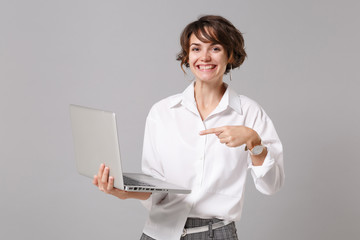 Wall Mural - Smiling young business woman in white shirt posing isolated on grey background in studio. Achievement career wealth business concept. Mock up copy space. Pointing index finger on laptop pc computer.