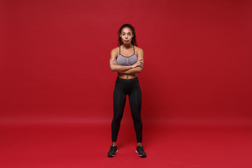 Confident young african american sports fitness woman in sportswear posing working out isolated on red background studio portrait. Sport exercises healthy lifestyle concept. Holding hands crossed.