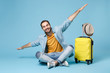 Smiling traveler tourist man in yellow clothes isolated on blue wall background. Male passenger traveling abroad on weekend. Air flight journey concept. Sit near suitcase, spreading hands like flying.