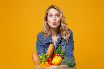 Wall Mural - Pretty young woman in denim clothes posing isolated on yellow orange background. Delivery service from shop or restaurant concept. Hold brown craft paper bag for takeaway mock up with food products.