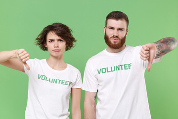 Displeased two young friends couple in white volunteer t-shirt isolated on pastel green background in studio. Voluntary free work assistance help charity grace teamwork concept. Showing thumbs down.