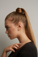 A blonde European girl with long straight hair in a high ponytail and a hair knot is posing on the gray background. The lady is wearing a black tee-shirt. 