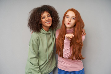 Wall Mural - Indoor photo of cheerful young lovely girlfriends hugging each other while posing over grey background in comfortable casual clothes, looking positively at camera