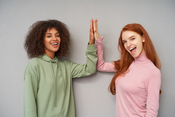 Wall Mural - Cheerful young attractive ladies raising their palms and giving five to each other while looking positively at camera with wide smiles, isolated over grey background