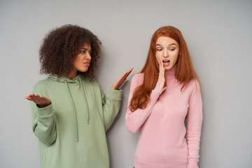 Wall Mural - Displeased young pretty curly dark skinned brunette female looking with dissapointed face at her redhead girlfriend and raisingmemotionally hands, isolated over grey background