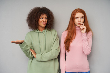Wall Mural - Dissatisfied young pretty dark skinned brunette woman in green hoodie raising confusedly her palm while looking at her lovely bewildered friend with long foxy hair, isolated over grey background