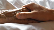 Worried Man Taking And Gently Stroking Hand Of His Sick Mother Showing Care Or Love. Son Comforting Wrinkled Arm Of Elderly Mom Lying At Bed. Guy Giving Support To His Old Parent. Side View Close Up