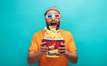 Handsome Emotional Young Guy In Orange T-shirt Eat Popcorn Wearing 3d Glasses Watch Film, Isolated On Blue Background.