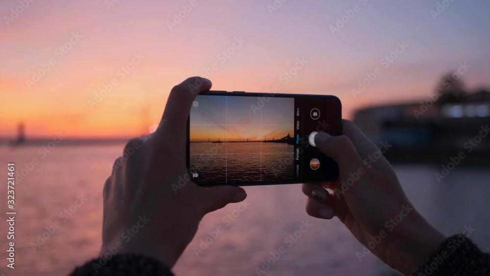 Close-up of a woman's hands with a phone taking photos of a bright sunset and the sea. - obrazy, fototapety, plakaty 