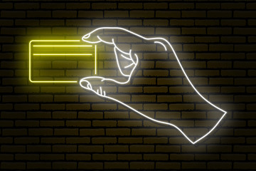 Neon sign. A human hand holding a gold plastic credit card with two fingers. Against a brick wall.