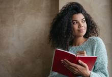 Beautiful African American Woman Holding Red Book, Looking At Window And Smiling. University Student Studying, Learning Language, Sitting At Library.  Portrait Of Young Pensive Writer Taking Notes