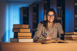 © bedya - Young happy smiling student in glasses preparing for the exam. Girl in the evening sits at a table in the library with a pile of books, smiling and looking at the camera