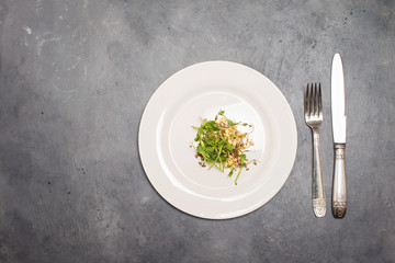 Fresh green sprouts microgreens and seeds in white plate on grey background with silverware and copy space.  Dietary nutrition. Small portion, weight loss concept.