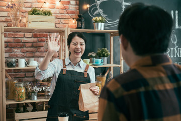 Wall Mural - startup small business takeaway food people and service concept. happy asian chinese woman seller giving paper bag say hello gesture to male customer at vegan cafe store.