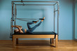 © Georgii - A young girl does Pilates exercises with a bed reformer, barrel machine tool. Beautiful slim fitness trainer on the background of a reformer doing various exercises gray, low key, light art. Fitness
