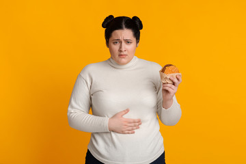 Stomachache. Girl Holding Burger And Touching Stomach Feeling Bad After Eating Junk Food Standing On Yellow Studio Background