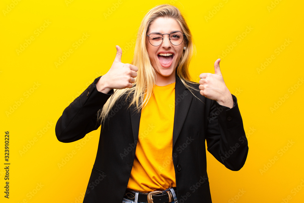 young pretty blonde woman smiling broadly looking happy, positive, confident and successful, with both thumbs up against yellow wall - obrazy, fototapety, plakaty 