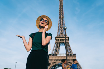 Wall Mural - Happy Caucasian female traveller enjoying smartphone conversation with friend during holidays time for visit romantic showplace, joyful woman in sunglasses and hat calling via mobile network app