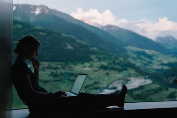 Wall Mural - Side view of skilled female digital nomad working remotely on freelance project sitting at table near big window with breathtaking view of high mountains,millennial woman typing content text on laptop