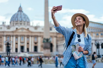 Wall Mural - Cheerful woman in joyful mood holding navigation map and clicking cellphone images via medial application on smartphone, happy Caucasian making video content while visiting St Peter Square in Vatican