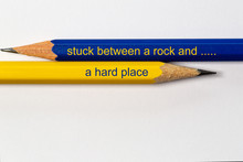 Two Sharpened Pencils In Blue And Yellow Point In Different Directions Isolated Against A White Background With The Conflicting Wisdom: Stuck Between A Rock And A Hard Place