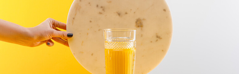 cropped view of woman holding marble circle near glass of fresh delicious yellow smoothie on grey and yellow background, panoramic shot