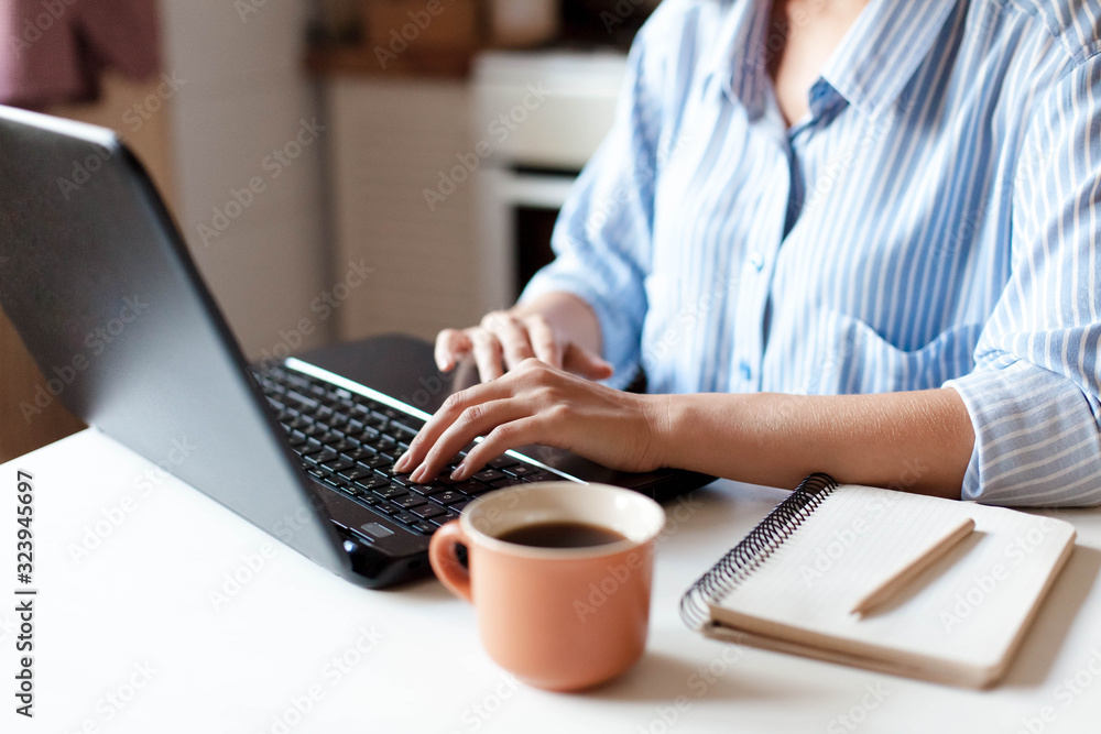 Freelancer workplace with laptop, cup of coffee and notebook. Young woman working from home office in cozy kitchen. Female business. Close up of hands. - obrazy, fototapety, plakaty 