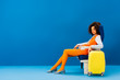 © LIGHTFIELD STUDIOS - african american in retro dress sitting on seat near travel bag on blue background