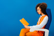 © LIGHTFIELD STUDIOS - side view of african american in retro dress sitting on seat and reading book isolated on blue