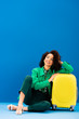 © LIGHTFIELD STUDIOS - smiling african american woman sitting near travel bag on blue background