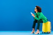 © LIGHTFIELD STUDIOS - african american woman sitting on travel bag and holding smartphone on blue background