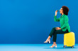 © LIGHTFIELD STUDIOS - side view of smiling african american woman sitting on travel bag and putting on sunglasses on blue background