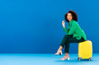 © LIGHTFIELD STUDIOS - smiling african american woman holding sunglasses and sitting on travel bag on blue background