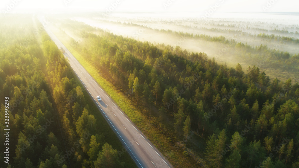 Aerial view of a highway with cars covered in fog. Early misty morning. Beautiful forest and sun rays.  Spotted from above with a drone. Finland, Europe. - obrazy, fototapety, plakaty 