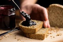 Person Applying Blueberry Jam On Fresh Slices Of Bread, Close-up Of The Moment When The Jam Touches The Bread, Warm Light, Wooden Table Top And Dark Background, Hands Visible