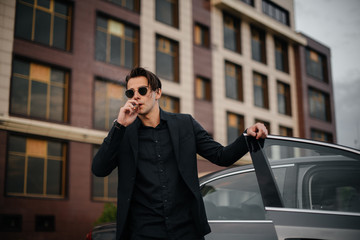 A stylish businessman smokes cigars near a luxury car. Fashion and business