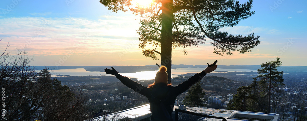 Obraz na płótnie Happy feeling with a view of the City of Oslo, Norway in front. Oslo fjord in the background. Women standing with her hands in the air. Sunbeams glittering through a tree. w salonie