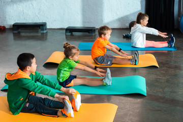 multicultural children stretching while sitting on fitness mats in gym