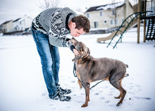 A Young Adult Latino Caucasian Man Wearing A Sweater And Blue Jeans Taking His Dog Out To Play In The Snow By The Neighborhood Playground.