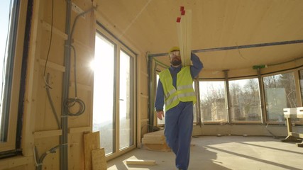 Wall Mural - SLOW MOTION, CLOSE UP, LOW ANGLE, LENS FLARE: Content worker wearing coveralls and hard hat arrives to work carrying wooden beams on his shoulder. Man carrying planks across house under construction.