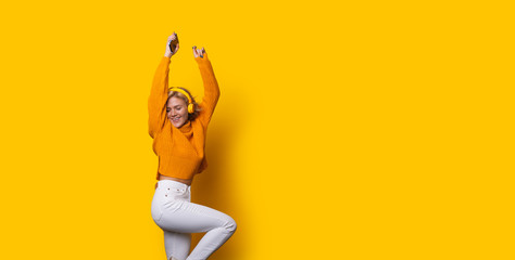 Stunning blonde caucasian girl dressed in an orange sweater and white jeans is dancing while posing on a yellow background and listening to music